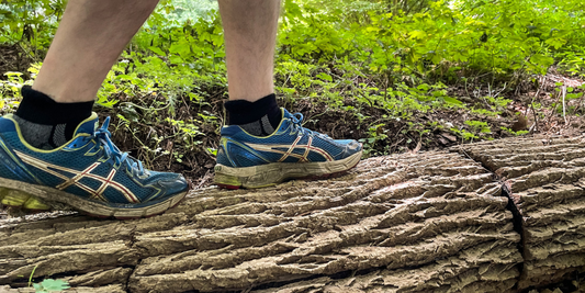 A person wearing alpaca ankle socks and blue sneakers walks on a log in the woods.