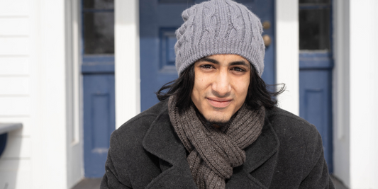 A guy wearing a grey knit alpaca beanie and ribbed charcoal alpaca scarf sits on a front porch with a blue door in the background