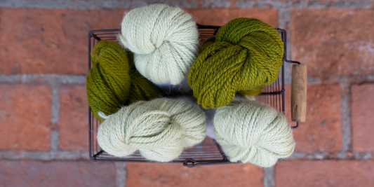 Deep green and cream colored alpaca yarn sits in a wire basket with a brick background