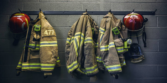 4 jackets of firefighters along with 2 helmets hang on a metal coat hanger in front of a cinder block wall.