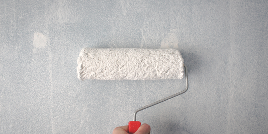 A roller with white paint on it is being pressed against a grey wall.