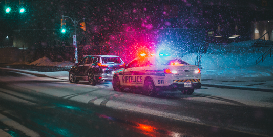 Photo shows a police car with its lights on pulling over a grey car at night.