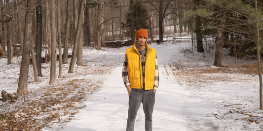 A guy wearing an alpaca hat and a yellow puffer vest stands on a snowy path in the woods