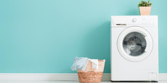 There is a white waching machine on the far right with a small potted plant on top and a laundry hamper on its left side that has clothes hanging over it. The wall behind it is light-medium blue with white trim.