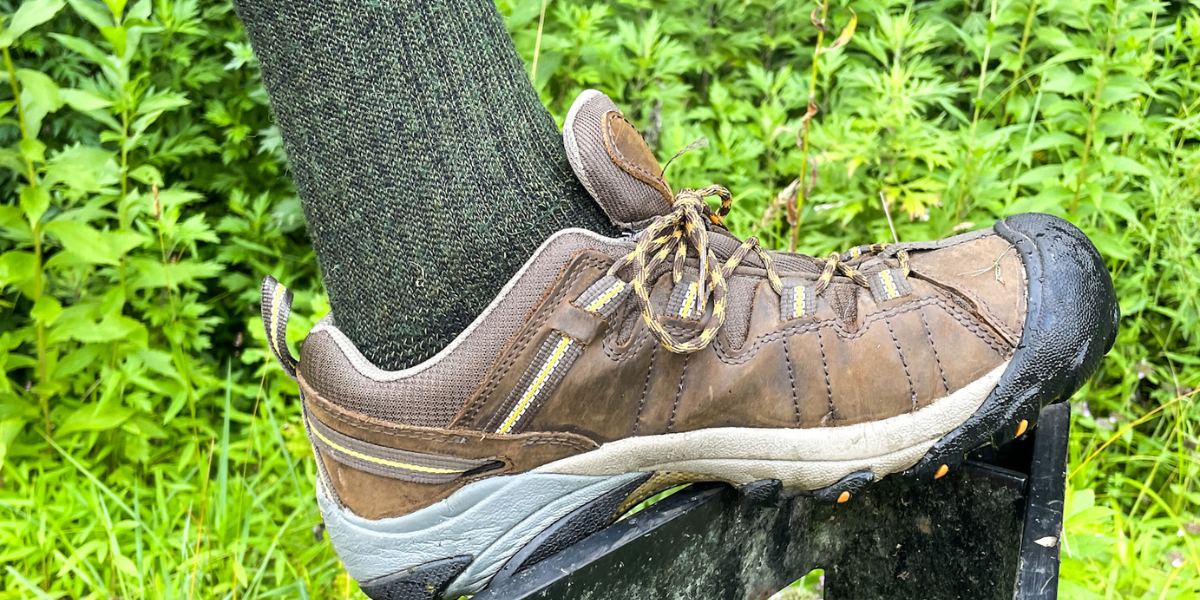 A person wearing hiking boots and a dark green alpaca boot sock rests their foot on a metal pole
