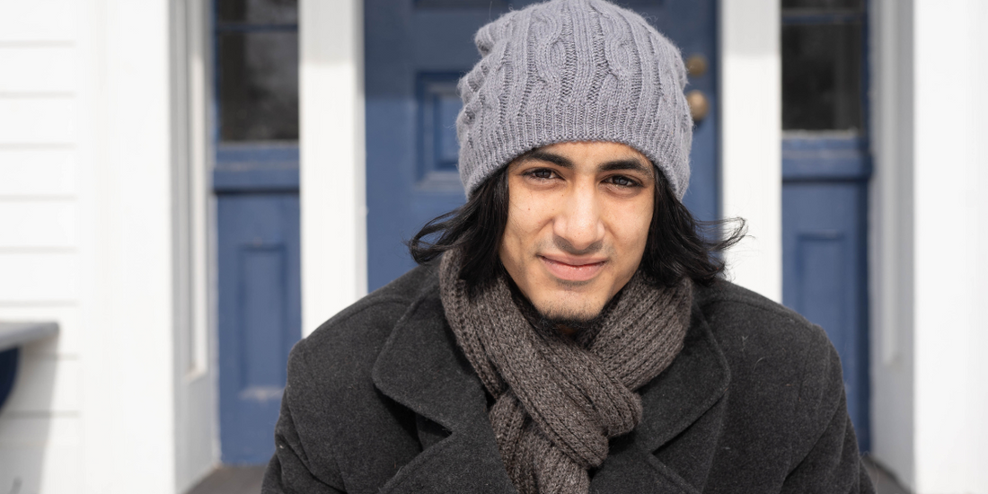 A guy wearing a grey knit alpaca beanie and ribbed charcoal alpaca scarf sits on a front porch with a blue door in the background