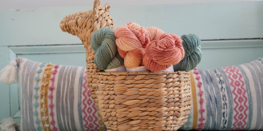 A woven basket shaped like a llama holds medium blue, peach, and pink skeins of yarn and is positioned in front of a multi-color long pillow on a light blue bench