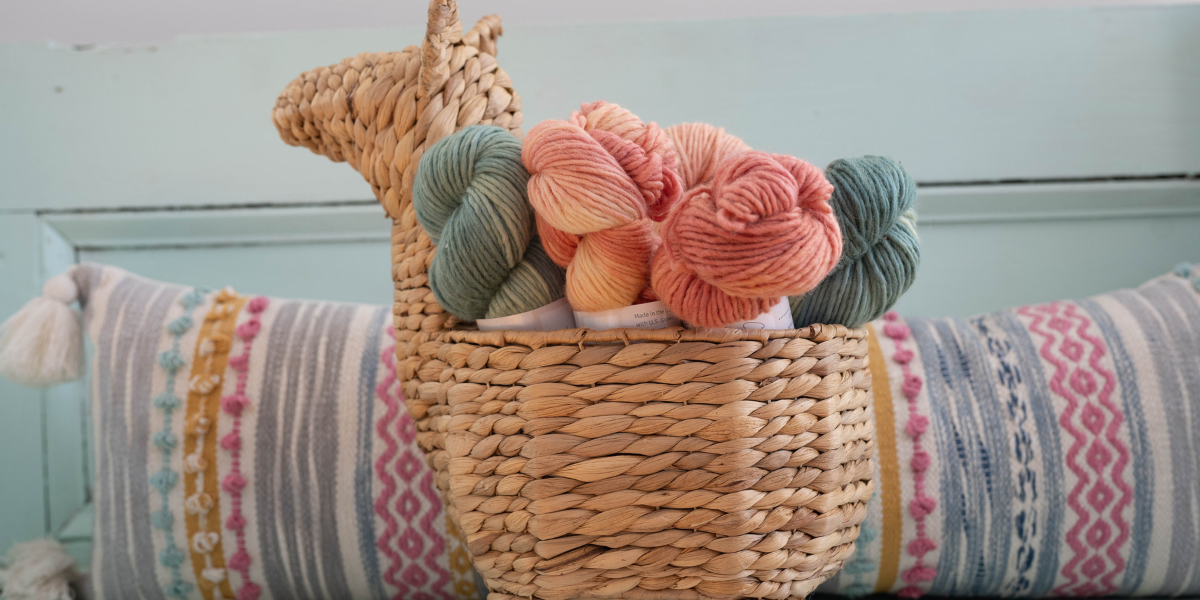 A woven basket shaped like a llama holds medium blue, peach, and pink skeins of yarn and is positioned in front of a multi-color long pillow on a light blue bench