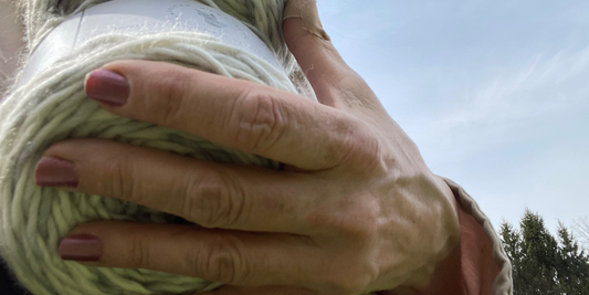 A hand grasps a cake of light green tinted yarn with the sky as the background