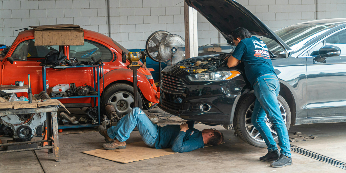 Two men work on a black car in a garage with a red volkswagen bug in the background