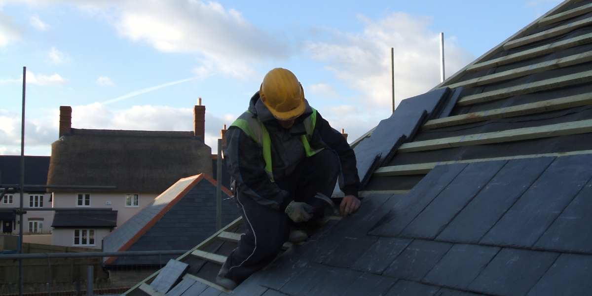 A roofer is laying tiles on a roof wearing a safety vest and hard hat.