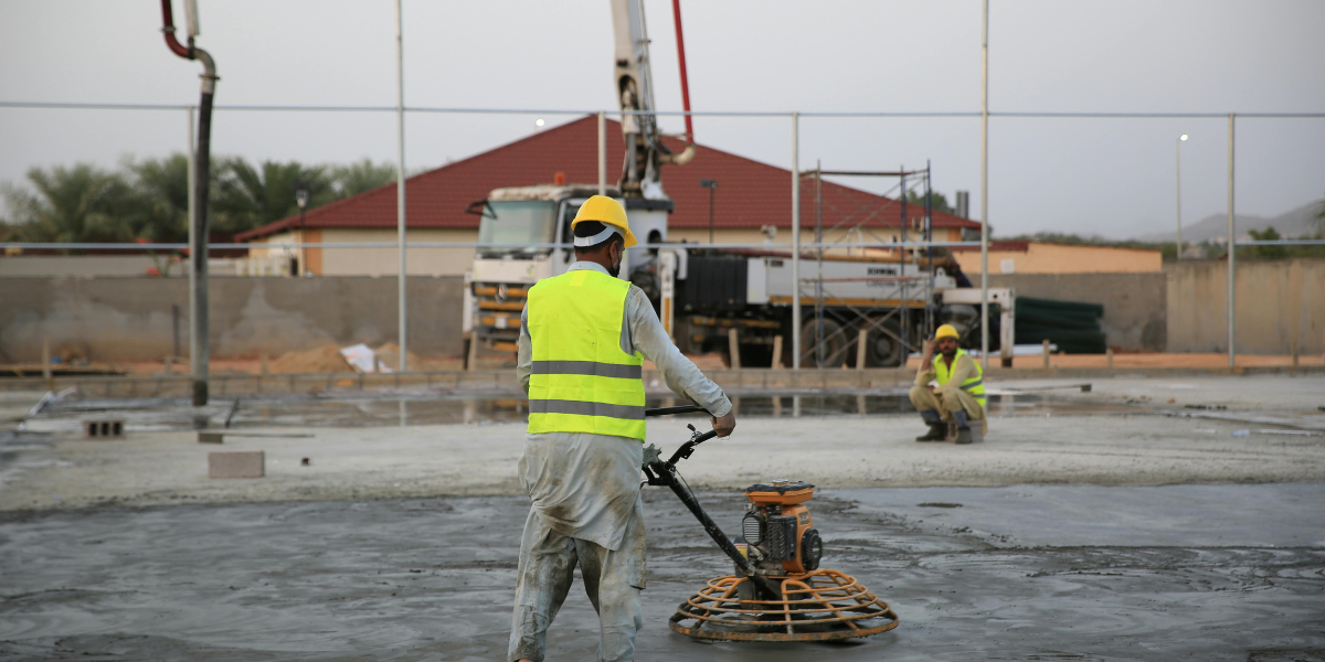 A person in a yellow safety vest and hard hat is smoothing concrete in an outdoor lot