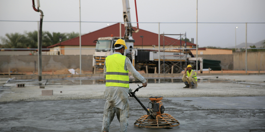 A person in a yellow safety vest and hard hat is smoothing concrete in an outdoor lot