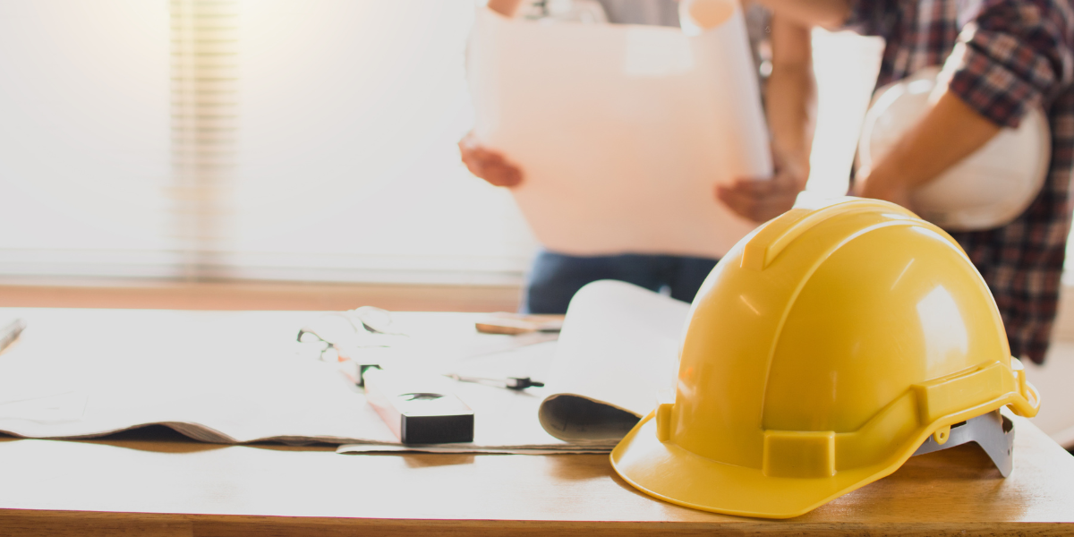 A table has construction blueprints and a hard hat on it with two people looking at plans in the background.