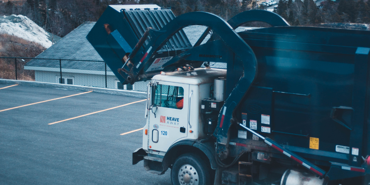 A garbage truck in a parking lot lifts a dumpster to dump in the back of the truck.