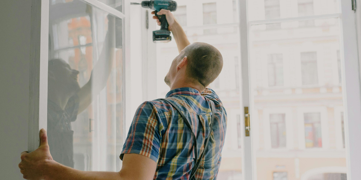 A man in a blue and yellow plaid shirt installs a window with a screw gun.