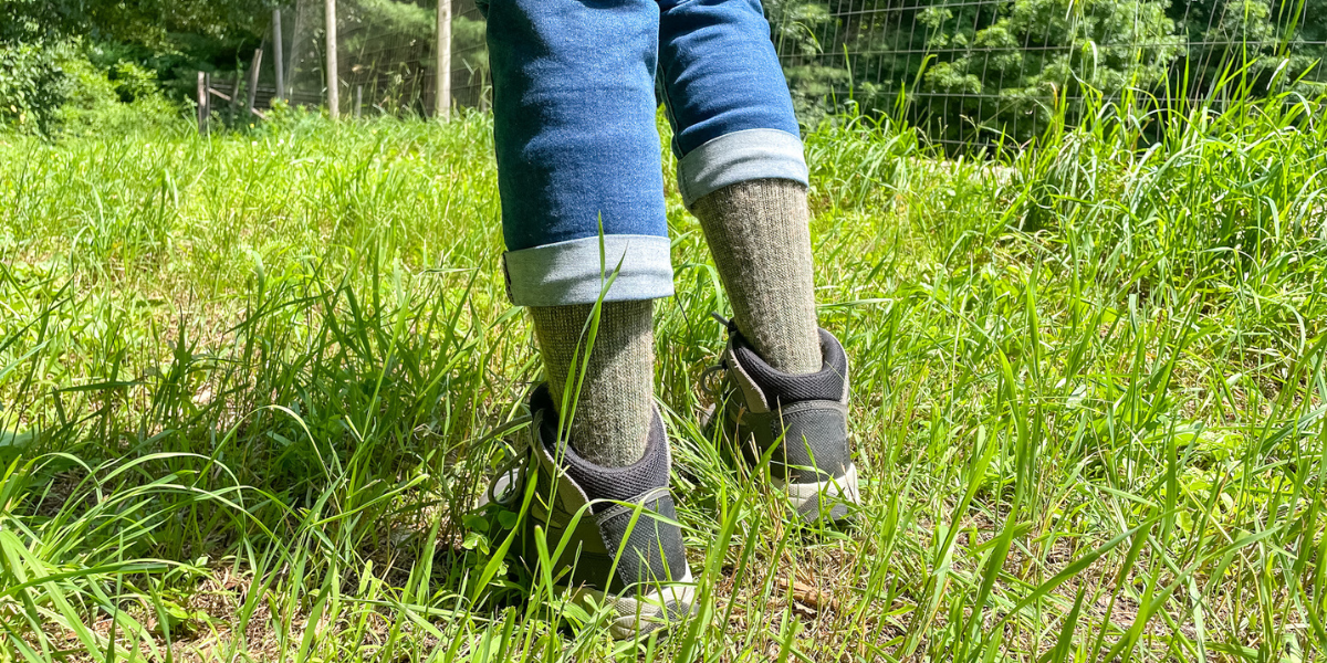A person wearing blue capris is standing in the grass wearing hiking boots and olive alpaca boot socks