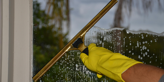 A person's hand is shown wearing a thick yellow glove and scraping soapy water off a window.