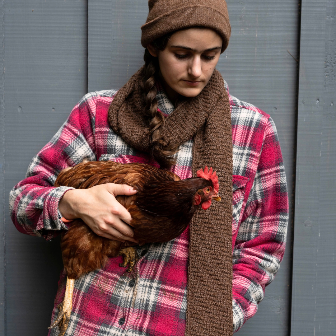 A woman wearing a brown knit beanie, brown scarf, and a red and white plaid flannel shirt holds a red chicken against a gray wooden background.