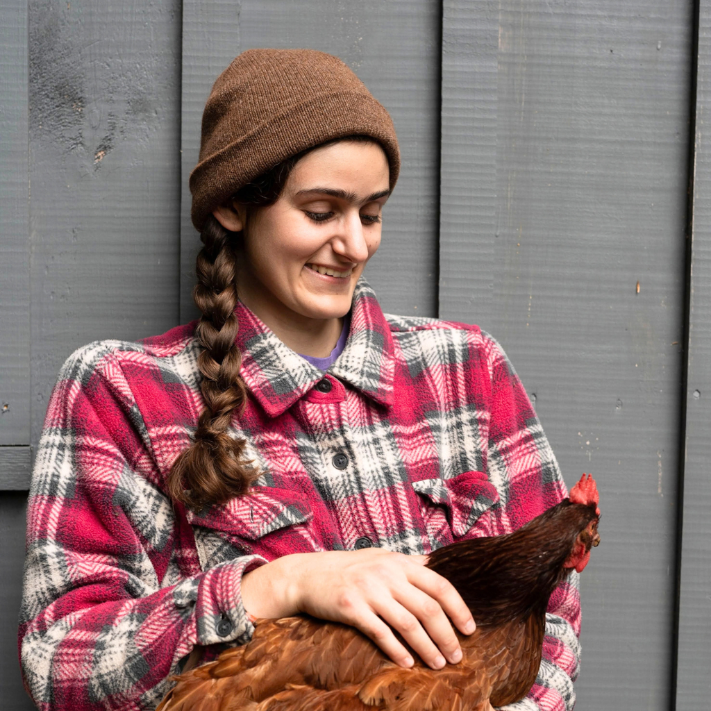 A woman wearing a brown knit beanie and a red, black, and white plaid flannel shirt holds a red chicken in front of a gray wooden wall.
