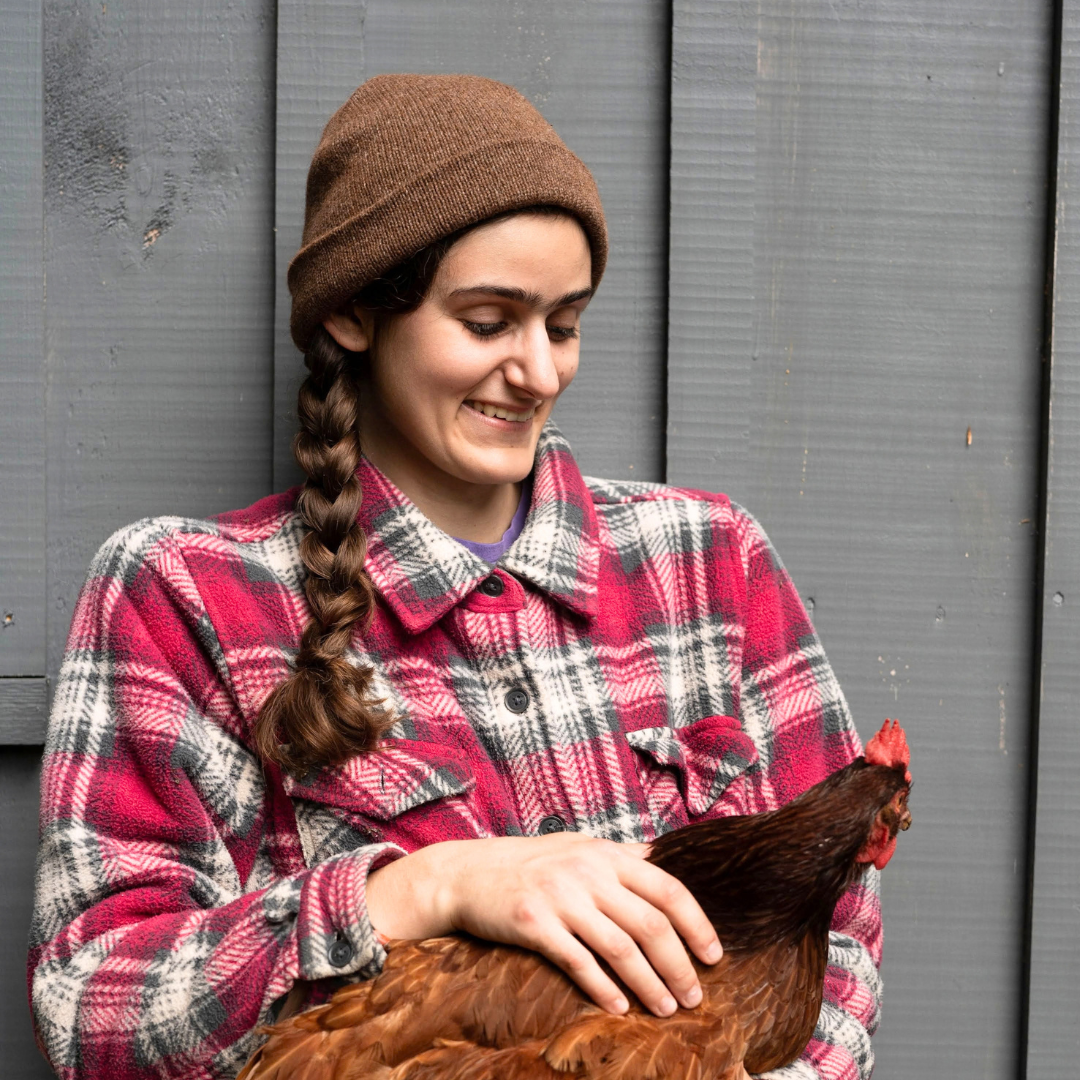 A woman wearing a brown knit beanie and a red, black, and white plaid flannel shirt holds a red chicken in front of a gray wooden wall.
