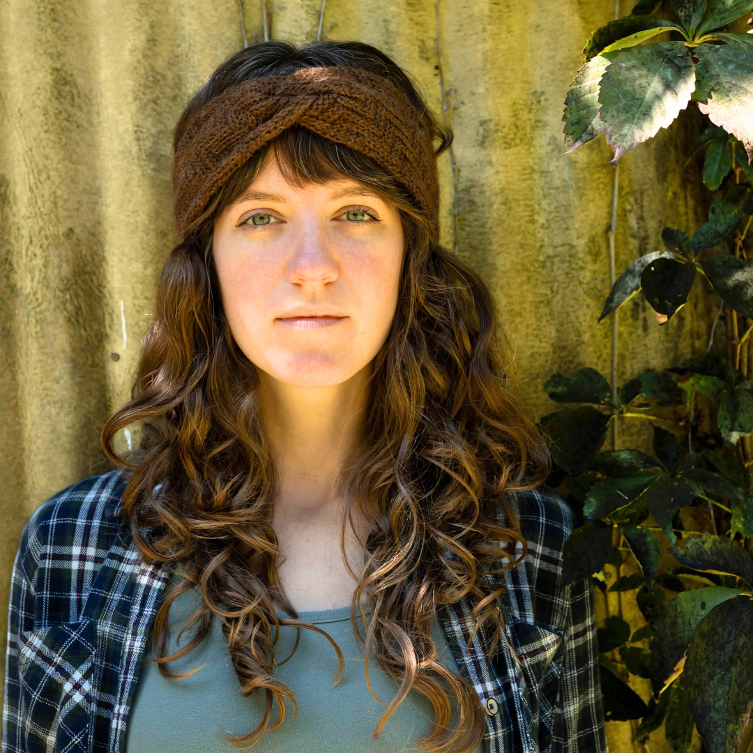A woman with long, wavy brown hair wearing a brown knitted twisted headband, a blue and white plaid flannel shirt, and a sage green top, standing in front of a corrugated metal wall with green plants on the right.