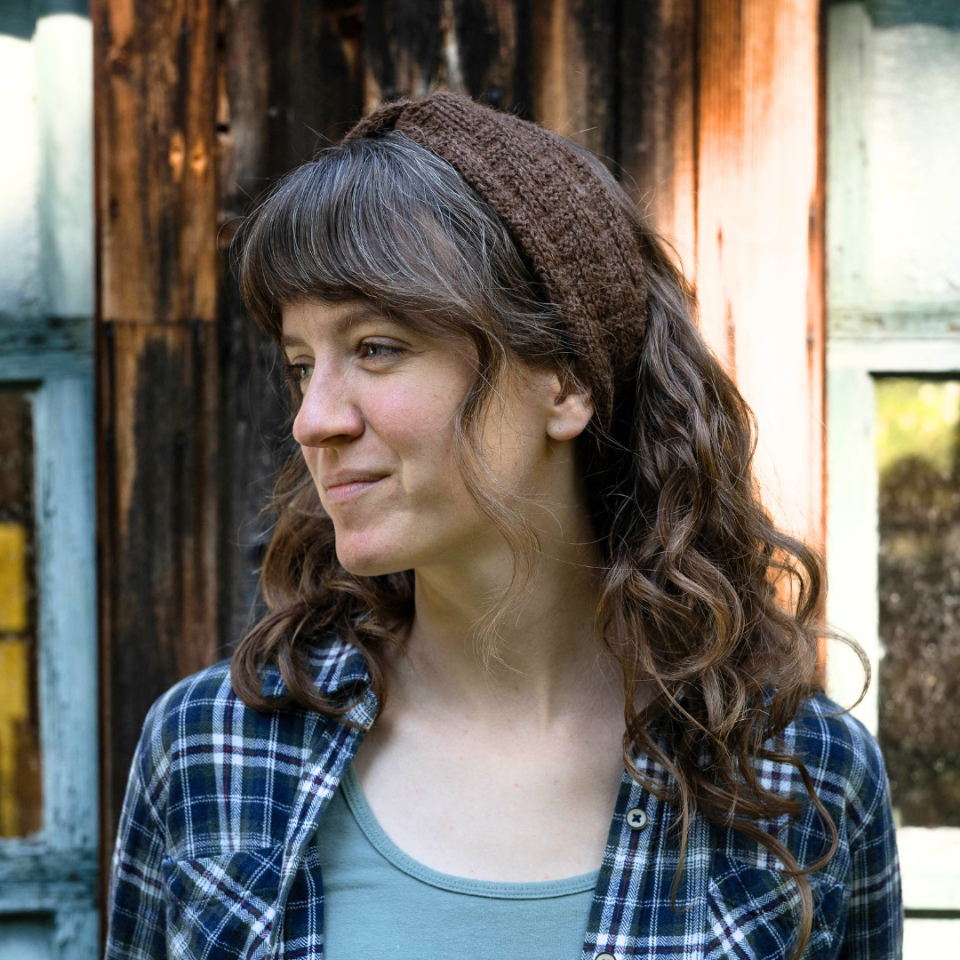 A woman with long brown curly hair wearing a brown knit headband, a blue and white plaid flannel shirt, and a sage green tank top, standing in front of a rustic wooden door.