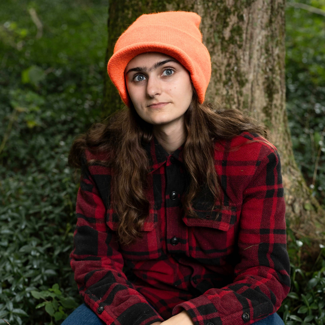 A young woman with long brown hair sits outdoors in front of a tree, wearing a bright orange knit beanie and a red and black buffalo plaid flannel shirt.