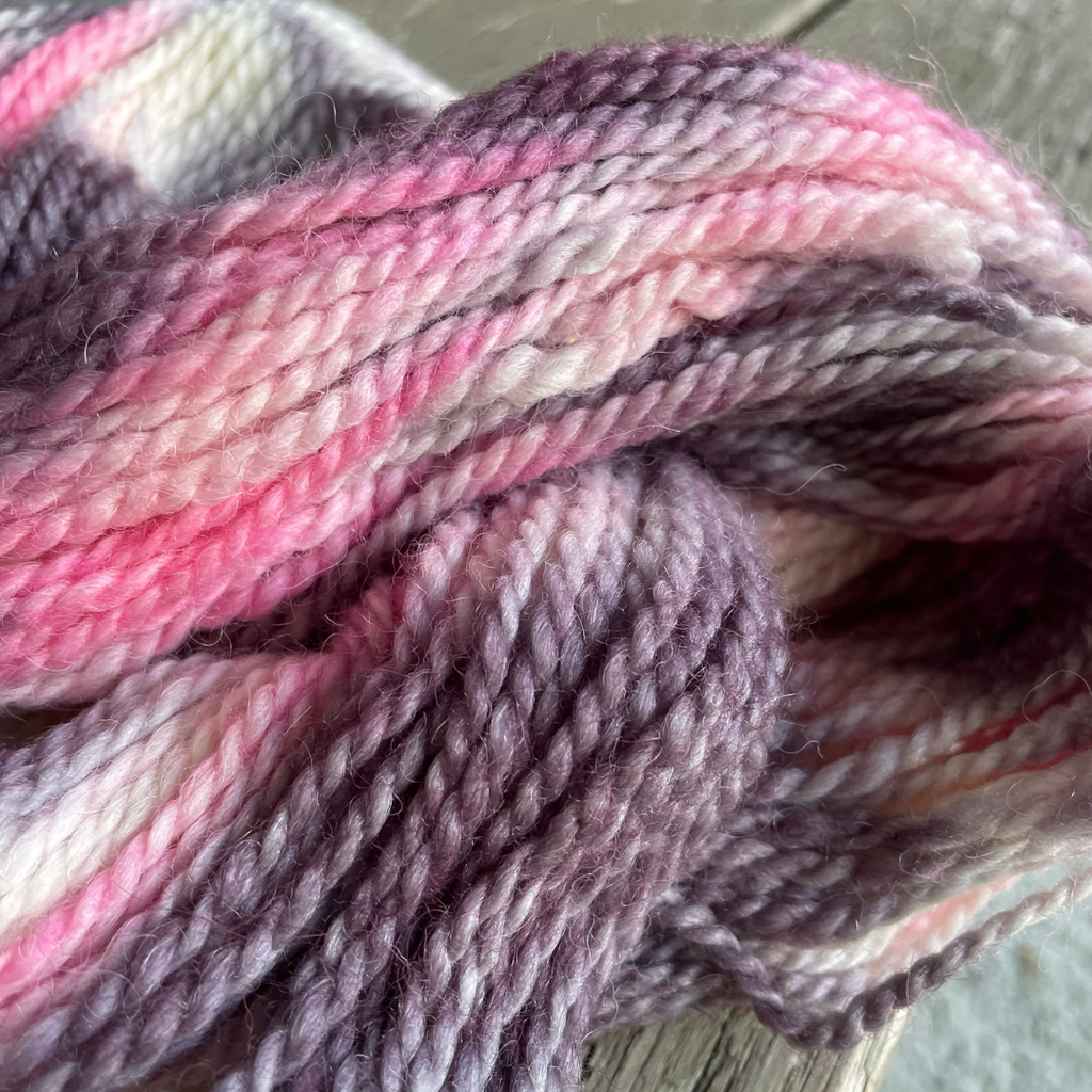 A close-up of variegated yarn with soft stripes of pink, mauve, lavender, cream, and muted brown, piled together on a light surface.