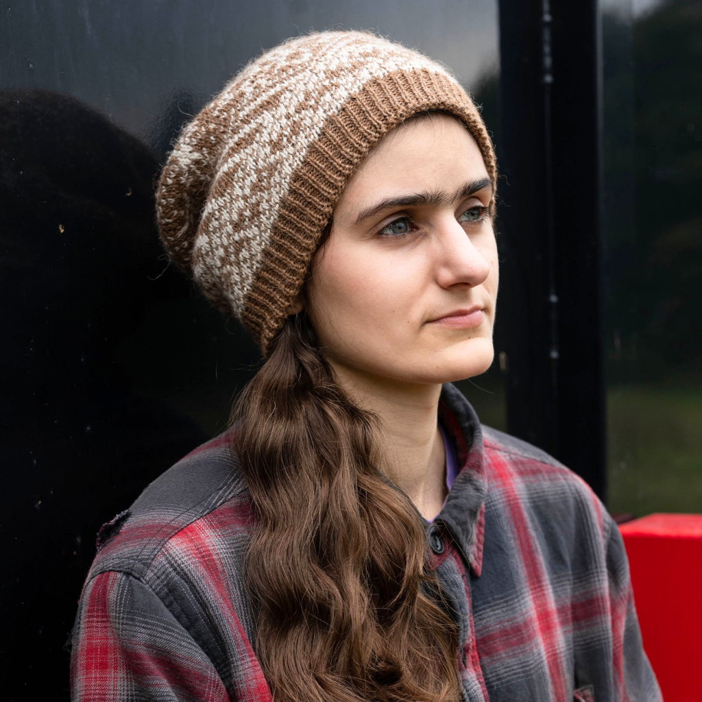 A woman wearing a brown and cream marled knit slouch beanie and a red and gray plaid flannel shirt, looking off to the side.