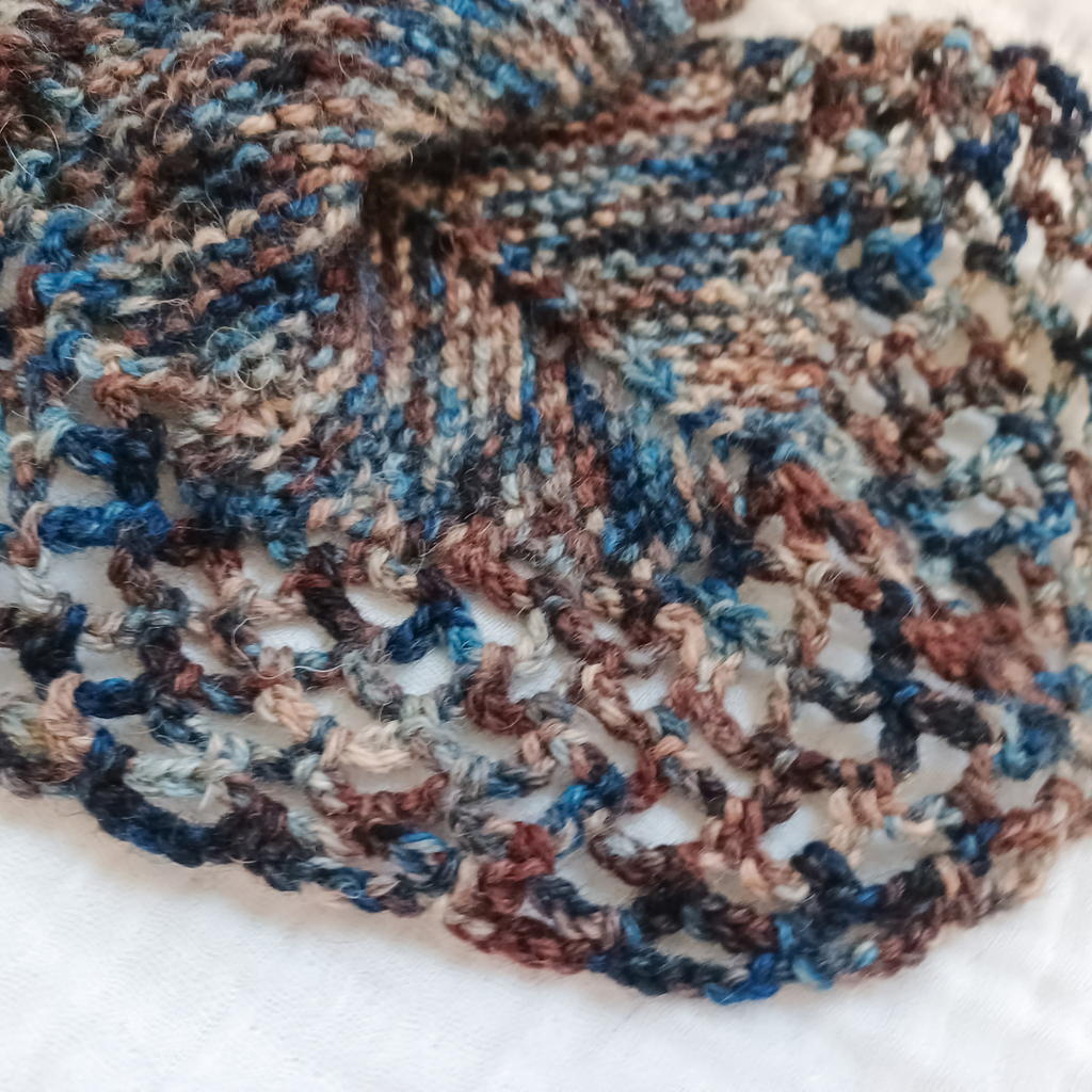 A close-up of a hand-knit lace cowl or scarf in variegated yarn with blue, brown, beige, and gray tones, laid flat on a white fabric background.